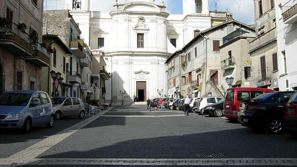 Piazza lastricata in un centro storico, con auto parcheggiate ai lati e palazzi residenziali che fiancheggiano la strada. Sullo sfondo si vede una grande chiesa barocca bianca con facciata simmetrica, portale centrale e due campanili laterali.