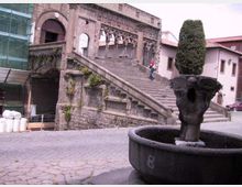 Scalinata in pietra di un edificio storico con archi decorati e portale, affacciata su una piazza lastricata. In primo piano si vede una grande fontana in pietra con un vaso centrale e una pianta potata, con impalcature sul lato sinistro dell’edificio.
