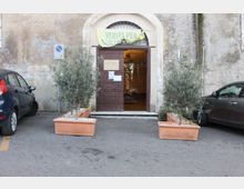 Ingresso di un edificio in muratura con intonaco consumato, portale ad arco e porta in legno aperta con luce interna visibile. Davanti all’entrata ci sono due grandi fioriere rettangolari con alberelli, affiancate da auto parcheggiate ai lati e un cartello stradale sulla parete.