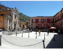 Piazza lastricata in un centro storico, circondata da edifici colorati con facciate tradizionali e finestre simmetriche, tra cui una chiesa in pietra con campanile. In primo piano ci sono paletti con catene che delimitano l’area pedonale, con colline verdi sullo sfondo.