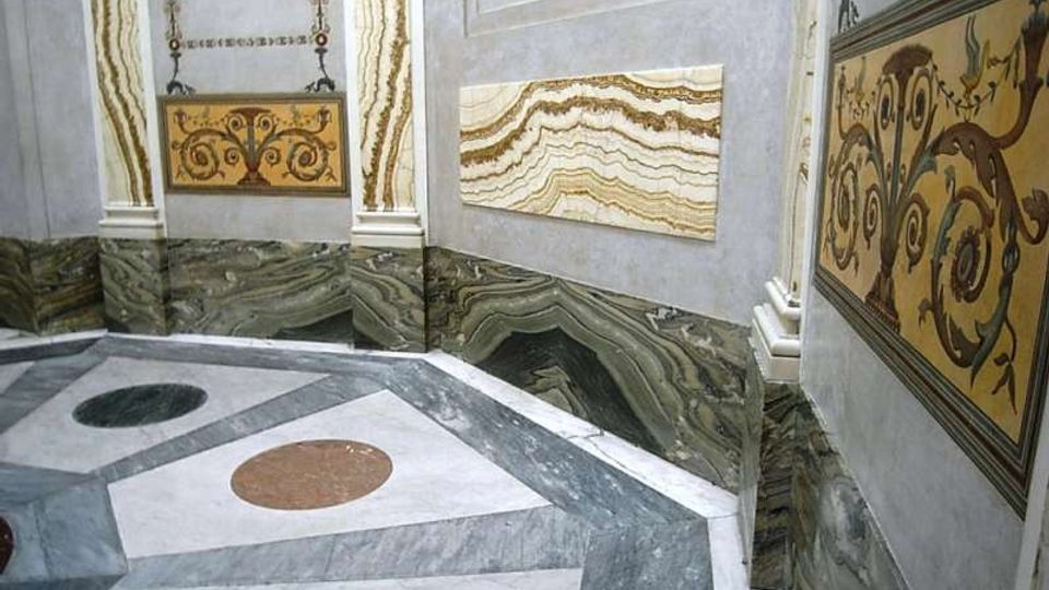 Interno decorato con pareti rivestite in marmo venato e pannelli ornamentali gialli con motivi a volute. Pavimento geometrico in marmo con riquadri e cerchi colorati, delimitato da zoccolature in pietra scura.