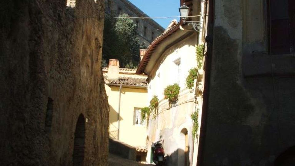 Vicolo stretto in salita con pavimentazione in pietra, fiancheggiato da alte mura in pietra e da edifici storici con intonaco chiaro e tetti in tegole. Sullo sfondo si vede una grande struttura fortificata in pietra sotto un cielo sereno.