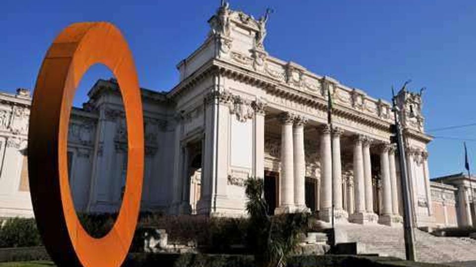 Facciata di un edificio monumentale in stile neoclassico con grande portico colonnato e scalinata in pietra, sotto un cielo sereno. In primo piano, su un prato, è visibile una grande scultura anulare arancione.