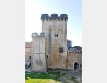 Torre medievale in pietra con merli e piccole finestre ad arco, affiancata da un bastione e da un muro di cinta con portale ad arco. Davanti si vede un’area di prato e terreno sterrato, con edifici in muratura sullo sfondo.