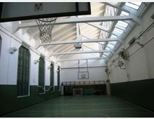Interno di una palestra scolastica con campo da basket, pareti bianche e verdi e grandi finestre ad arco su un lato. Il soffitto alto con travi a vista e lucernari lungo la falda illumina lo spazio; si vedono canestri e attrezzature sportive sul fondo.