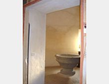 Interno di una piccola stanza con pareti intonacate chiare e pavimento in mattonelle, vista attraverso una porta. Al centro si trova una grande vasca circolare in pietra su base, illuminata da una lampada sul lato destro.