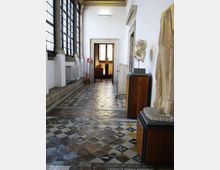 Corridoio interno di museo o palazzo storico con grandi finestre a griglia sul lato sinistro e pavimento a mosaico geometrico multicolore. Sulla destra sono presenti piedistalli con sculture e un passaggio con cornice in pietra che conduce a una sala sullo sfondo.