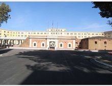 Ampio piazzale asfaltato davanti a un complesso architettonico con ingresso centrale in mattoni e pietra, con cancello ad arco e finestre piccole. Sullo sfondo si vede un grande edificio istituzionale giallo a più piani, con facciata simmetrica e una bandiera sul tetto.