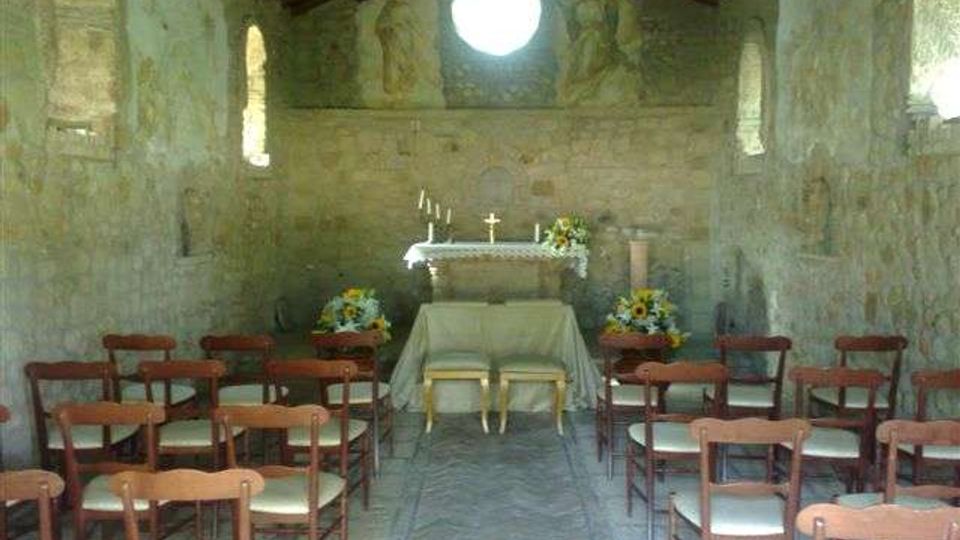 Interno di una piccola cappella in pietra con soffitto a travi in legno, navata centrale con sedie in legno e altare rivestito da tovaglia bianca. Sul fondo si vede un grande oculo circolare sopra l’altare, con affreschi sbiaditi e strette finestre laterali.