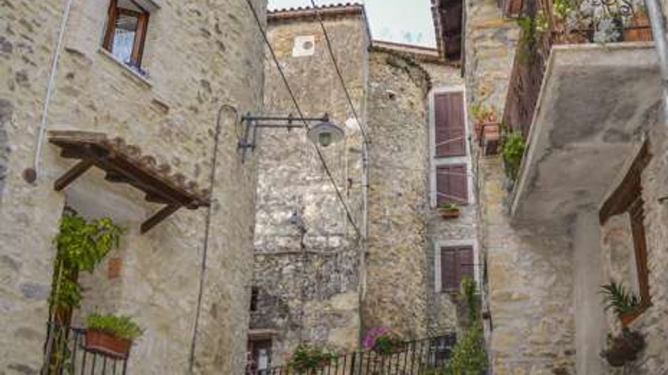Vicolo stretto in pietra con pavimentazione in ciottoli in un borgo storico, circondato da edifici in muratura con balconi, piccole finestre e gronde in legno. Sono visibili ringhiere, vasi con piante e una lampada a muro lungo la salita.