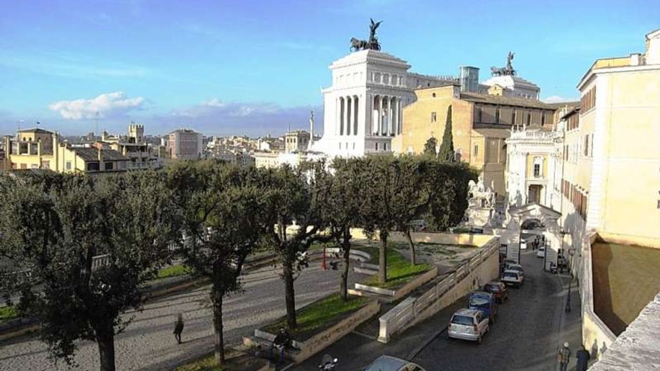 Veduta urbana con una strada in salita costeggiata da auto parcheggiate e alberi, affiancata da una terrazza con muretti e sculture. Sullo sfondo si distingue un grande edificio monumentale bianco con colonne e quadrighe in cima, circondato da palazzi storici.