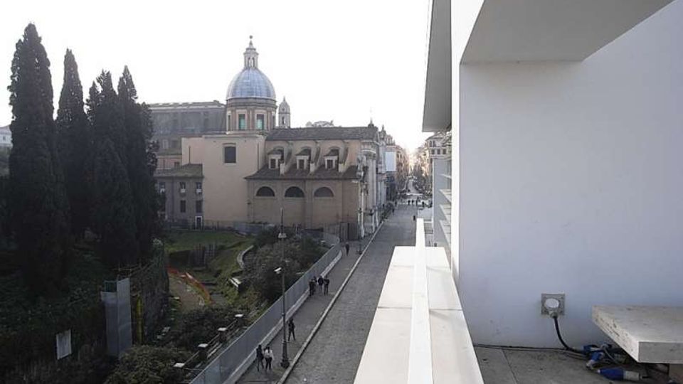 Vista urbana da una terrazza con parapetto bianco su una strada pedonale sottostante; sullo sfondo si vede un edificio storico con cupola e facciate ad archi. A sinistra c’è un’area verde recintata con alti alberi, mentre a destra si notano pareti e sporgenze di un edificio moderno.