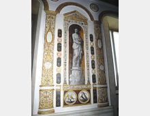 Parete interna decorata in stile classico con pilastri riccamente dorati, archi e medaglioni ornamentali. Al centro, una nicchia con statua dipinta in monocromo incorniciata da fregi, e sotto due pannelli circolari con uccelli, accanto a una finestra laterale.