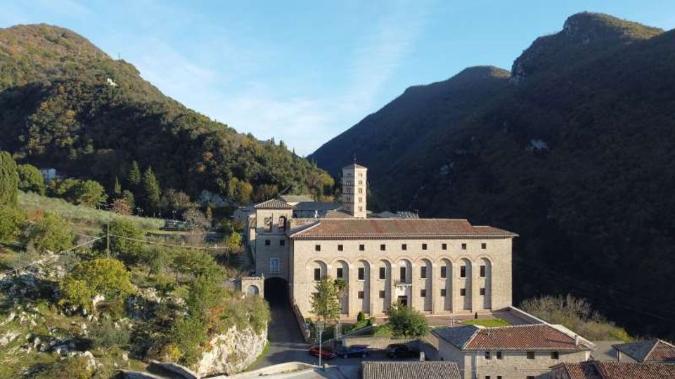 Grande edificio in pietra con tetto in coppi rossi e alte finestre ad arco, con una torre campanaria centrale, situato in una valle tra colline boscose. Intorno si vedono strade, piccoli edifici e vegetazione su pendii rocciosi sotto un cielo sereno.