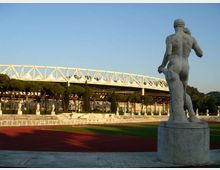Statua in pietra su piedistallo accanto a una pista di atletica, con sullo sfondo un grande stadio con struttura bianca a traliccio e alberi allineati lungo le gradinate sotto un cielo sereno.
