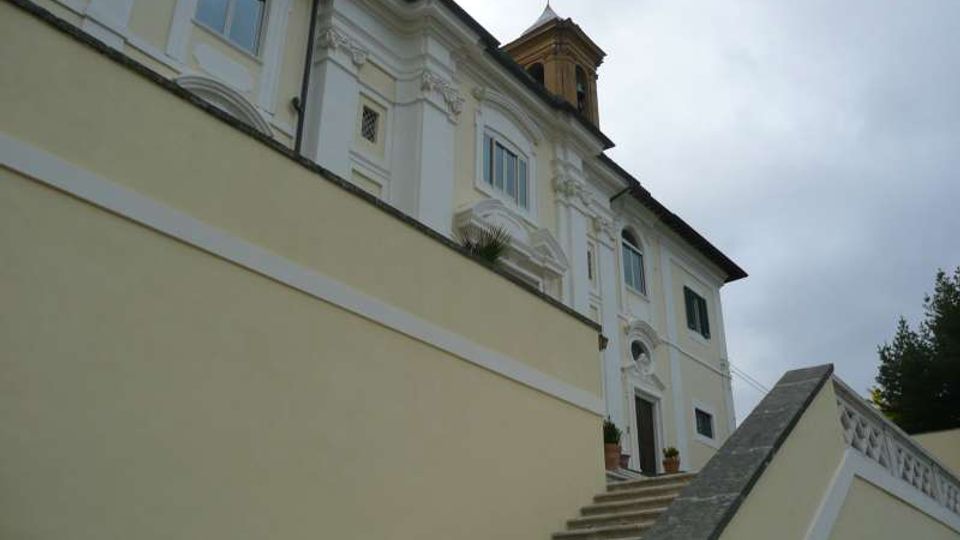 Facciata di un edificio storico chiaro con decorazioni in stucco e finestre ad arco, vista dal basso lungo una scalinata esterna con parapetto. Sul tetto si intravede una piccola torretta e sullo sfondo c’è cielo coperto con vegetazione laterale.
