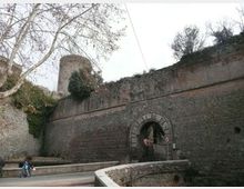 Mura di fortificazione in mattoni con grande arco d’ingresso in pietra e passaggio sottostante, affiancate da un torrione cilindrico sullo sfondo. Davanti si vede una strada con muretto curvo e alberi spogli lungo il lato sinistro.