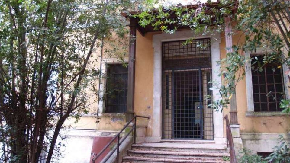 Ingresso di un edificio in muratura con intonaco color ocra, raggiunto da una scalinata con corrimano e coperto da una piccola tettoia. La porta è protetta da una grata metallica e la facciata è parzialmente nascosta da alberi e vegetazione fitta.