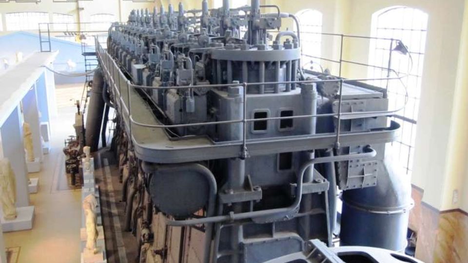 Grande sala industriale con ampie finestre ad arco, che ospita un enorme macchinario metallico grigio con tubazioni, valvole e passerelle con corrimano. L’ambiente è illuminato dalla luce naturale e presenta pavimento in legno e strutture portanti a vista.