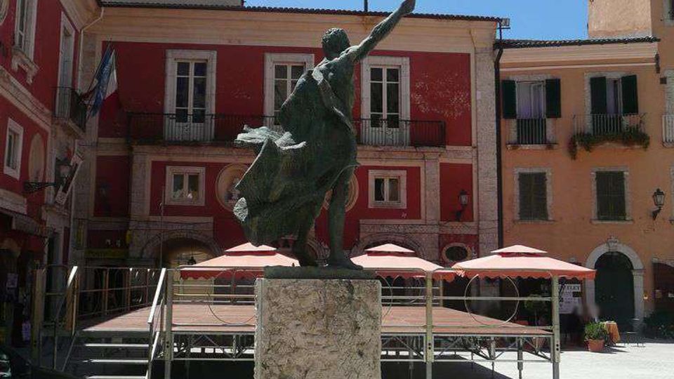 Statua in bronzo su alto piedistallo di pietra al centro di una piazza lastricata, con palco temporaneo e ombrelloni sullo sfondo. Intorno si vedono edifici a più piani con facciate colorate, balconi e finestre affacciate sulla piazza.