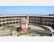 Cortile interno di una grande struttura circolare in muratura, con due livelli di porticati ad archi e finestre lungo l’anello esterno. Al centro si trova un piccolo edificio ottagonale con tetto a cupola, circondato da pavimentazione in cemento e aree con vegetazione spontanea.