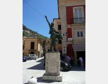Statua in bronzo su alto basamento in pietra al centro di una piccola piazza, circondata da edifici storici con facciate colorate e balconi. Sullo sfondo si vede una strada con auto parcheggiate e una collina verde sotto cielo sereno.
