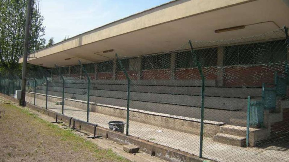 Tribuna di un impianto sportivo all’aperto con gradinate in cemento sotto una lunga copertura piatta, vista dietro una recinzione metallica. Nell’area circostante si notano un palo alto e alberi sullo sfondo.