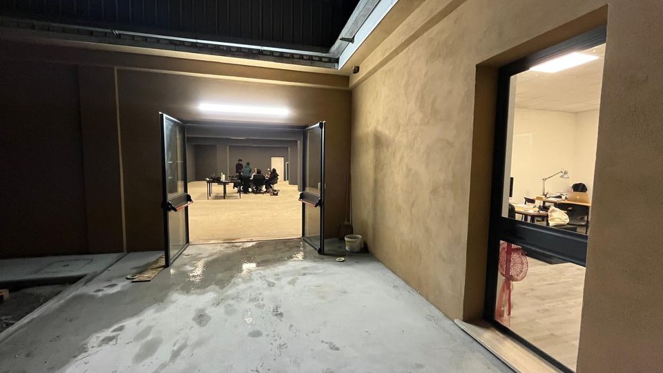 Cortile interno di un edificio moderno con pavimento in cemento e pareti color sabbia, illuminato da una lunga lampada a soffitto e con porte a doppia anta aperte verso un grande spazio interno. A destra una finestra mostra un ufficio con scrivanie e lampade da lavoro.