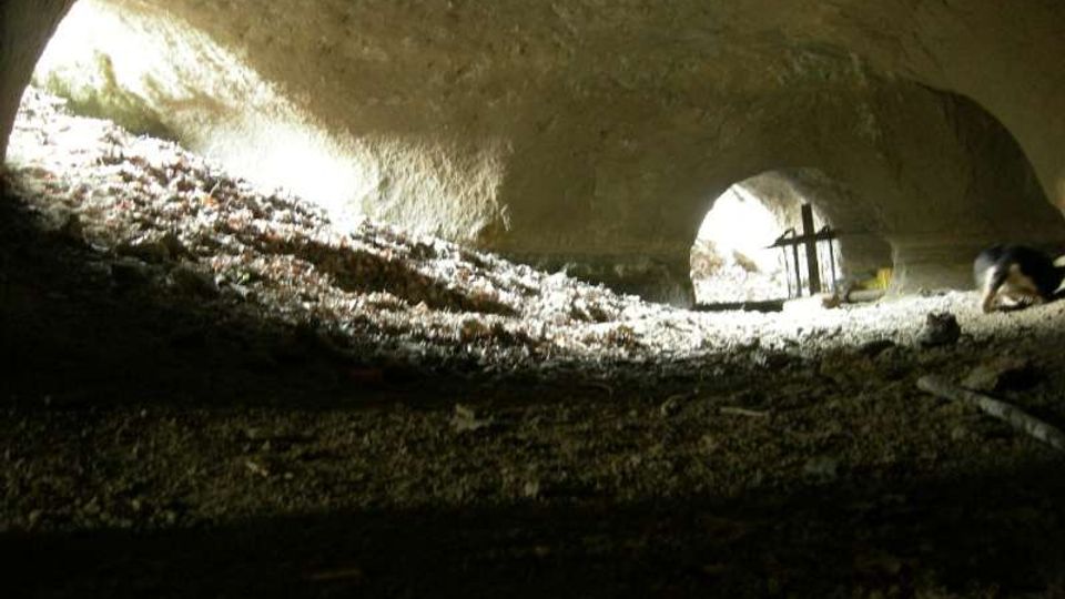 Interno di una grotta naturale con soffitto roccioso basso e irregolare, illuminata da aperture laterali che lasciano entrare fasci di luce. Il pavimento è ricoperto di terra e detriti, con un piccolo tavolo e oggetti visibili in lontananza vicino all’uscita.