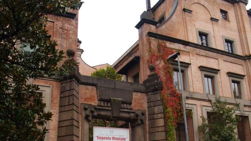 Edificio storico in mattoni con facciata alta e finestre incorniciate, con elementi architettonici scuri e un piccolo torrione sul tetto. In primo piano si vede un ingresso monumentale con pilastri e cancello, un’insegna, vegetazione rampicante e auto parcheggiate lungo la strada.
