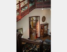 Interno di una casa storica con scala in legno e ringhiera in ferro battuto decorato, affacciata su un pianerottolo con parquet e tappeti. Sulla parete ci sono una vetrina in legno con oggetti esposti, un quadro ovale e una porta a vetri che conduce a un corridoio con colonne e tende.