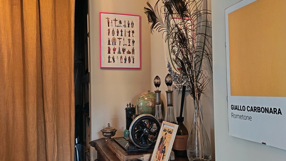 Angolo di un interno con pareti beige, una lanterna a soffitto e una tenda pesante che copre un’apertura. Accanto, un mobile in legno scuro con oggetti decorativi e un vaso con piume di pavone, mentre sulla parete sono appesi riquadri e pannelli con campioni di colore.