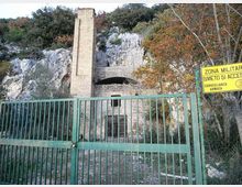 Cancello metallico verde che chiude l’accesso a una struttura in pietra incassata nella parete rocciosa, con alto camino in mattoni e portone murato. A destra è visibile un cartello giallo “Zona militare, divieto di accesso”, circondato da vegetazione e alberi.
