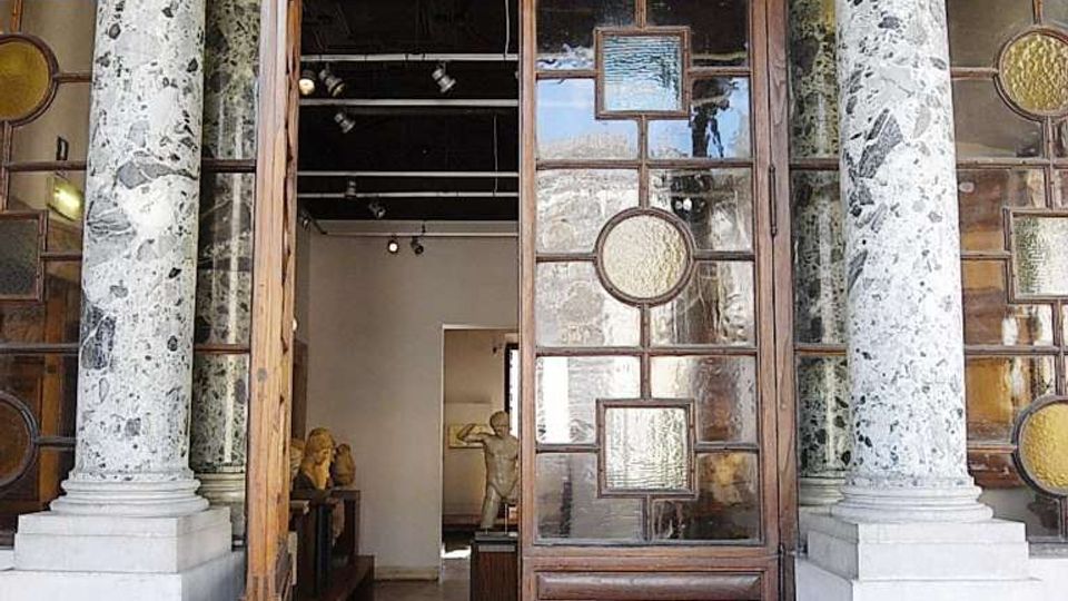 Ingresso di un edificio storico con due colonne in marmo maculato e capitelli decorati, che incorniciano una grande porta in legno con pannelli di vetro geometrici. Attraverso la porta aperta si intravede un interno tipo galleria o museo con pavimento scuro, illuminazione a soffitto e sculture esposte.