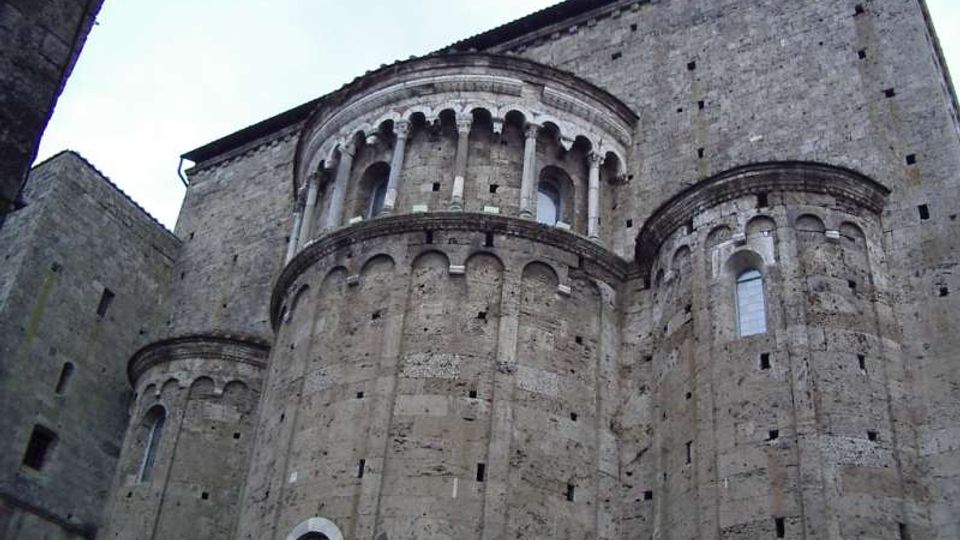 Vista esterna di una chiesa romanica in pietra con absidi semicircolari e decorazioni ad archetti ciechi. La facciata laterale mostra un alto tamburo con loggiato ad arcate e piccole finestre, su muratura massiccia in blocchi di pietra.