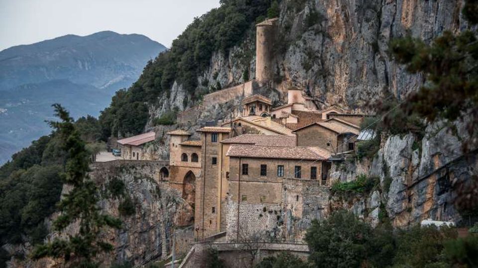 Monastero in pietra con tetti in tegole rosse costruito su una parete rocciosa a strapiombo, con torre cilindrica e arcate. Intorno si vedono scogliere e boschi di montagna, con vette sullo sfondo.