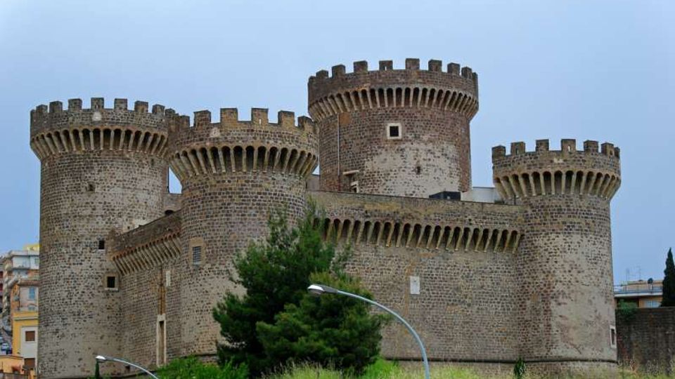 Castello medievale in pietra con quattro torri cilindriche merlate e bastioni, visto dall’esterno. In primo piano ci sono un’area verde con alberi e lampioni, con edifici urbani sullo sfondo sotto un cielo nuvoloso.