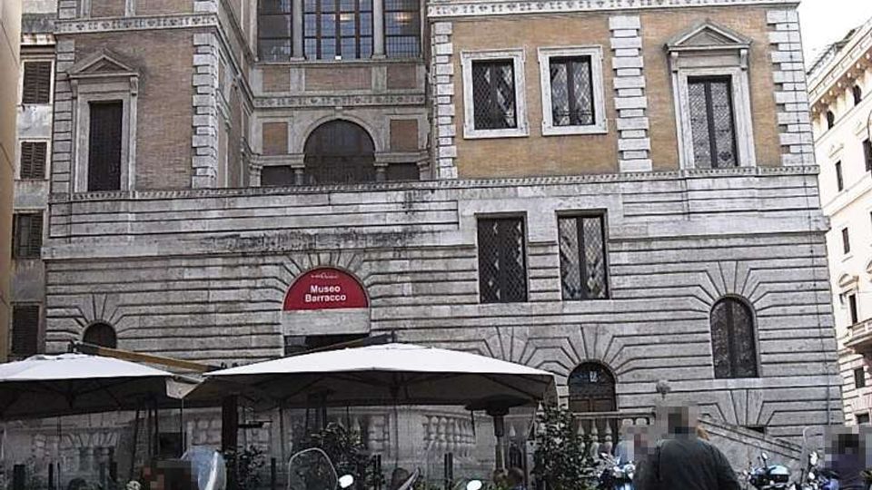 Facciata di un edificio storico in pietra e mattoni, con finestre rettangolari e cornicioni, identificato da un’insegna “Museo Barocco” sopra un ingresso ad arco. In primo piano si vede una strada urbana con molte moto parcheggiate e ombrelloni di un’area esterna.