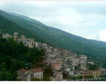 Borgo di montagna con case in pietra addossate al pendio, dominato da una chiesa e un campanile. Sullo sfondo si vedono colline boscose e montagne sotto un cielo nuvoloso.
