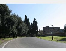 Strada asfaltata ampia e curva che attraversa un’area verde, fiancheggiata da alberi alti e lampioni. Sullo sfondo si vede un edificio in muratura con un muro di recinzione, sotto un cielo limpido.
