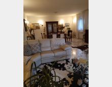 Soggiorno ampio con pavimento in marmo chiaro, divano ad angolo beige con cuscini decorativi e tappeti in stile pelle bovina. Sullo sfondo zona pranzo con tavolo e sedie, credenza in legno scuro e pareti con quadri e applique, accanto a una finestra con tende bianche.