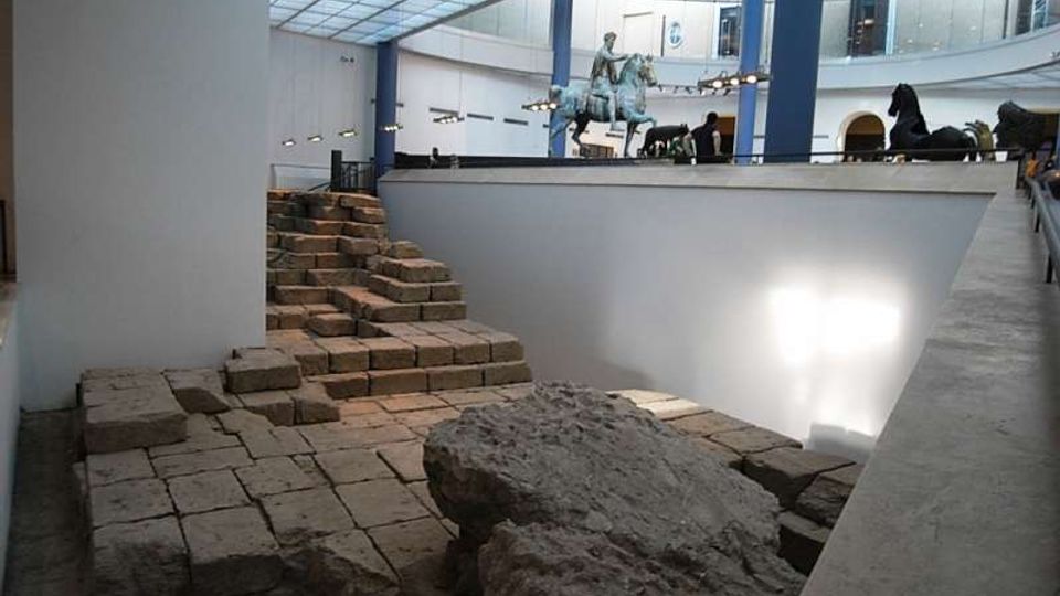 Interno di un museo con copertura in vetro e colonne, affacciato su un’area archeologica ribassata con pavimentazione in pietra, gradini e grandi blocchi rocciosi. Su una piattaforma superiore si intravedono sculture, tra cui una statua equestre, lungo il bordo del vuoto centrale.