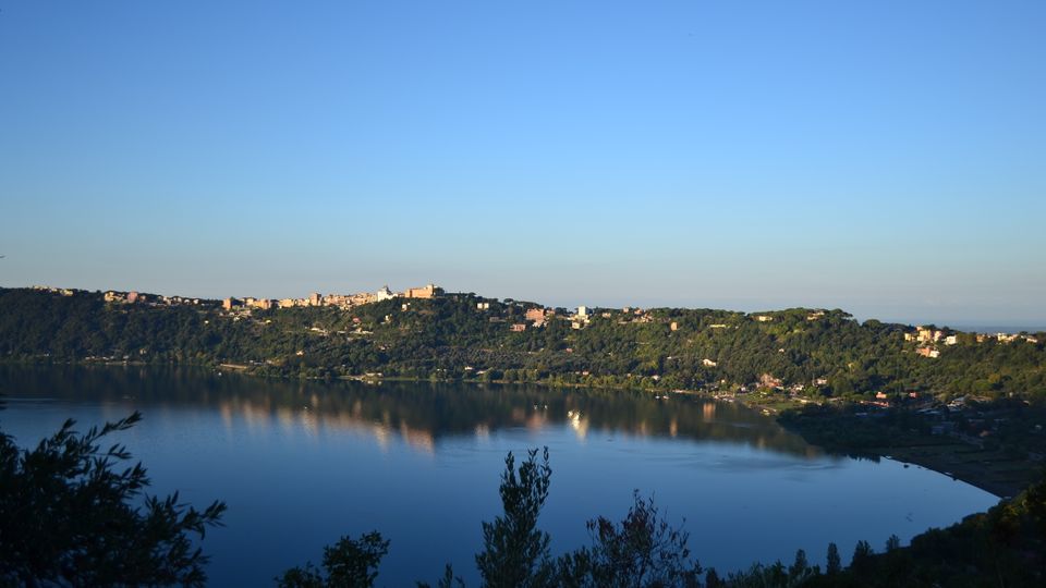 Vista panoramica di un lago calmo con riflessi sull’acqua, circondato da colline boscose. Sulla cresta delle colline si vedono abitazioni e piccoli edifici, sotto un ampio cielo sereno.