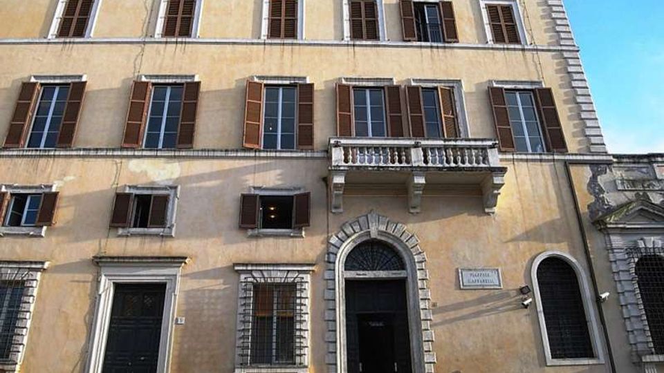 Facciata di un edificio storico a più piani con intonaco chiaro, finestre rettangolari con scuri in legno e un piccolo balcone in pietra sopra un portale ad arco con cornice bugnata. Al piano strada si vedono grandi porte scure e finestre con inferriate, con cielo azzurro sullo sfondo.
