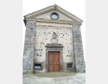 Facciata in pietra di una piccola chiesa o cappella storica con timpano triangolare e oculo circolare in alto. Al centro si vede un grande portale in legno incorniciato da pietra, con stemma decorativo sopra e due piccole finestre con inferriate ai lati.