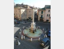 Fontana monumentale al centro di una piazza lastricata, con grande vasca circolare e colonna in pietra decorata con sculture. Intorno si vedono palazzi storici dai toni pastello con tetti in tegole e una strada stretta che si apre tra gli edifici.