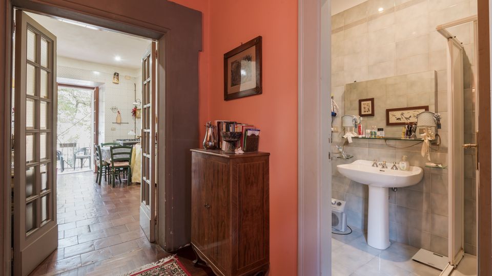 Corridoio interno con pareti rosa e pavimento in legno, con mobile in legno e porta a vetri che conduce a una cucina-sala da pranzo con accesso a un balcone. Sulla destra si apre un bagno piastrellato in beige con lavabo su colonna, specchio e box doccia.