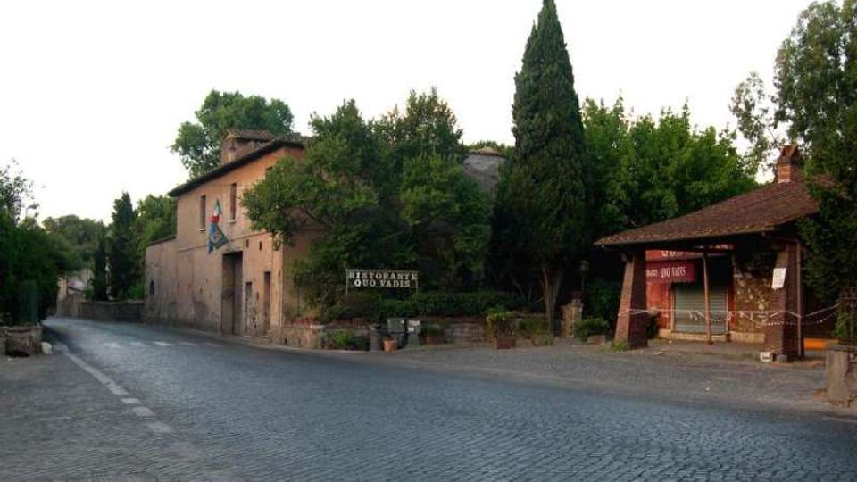 Strada lastricata in pietra in un’area rurale, con un edificio in muratura stile cascina e vegetazione alta sullo sfondo. A destra si vede una piccola pensilina in legno e mattoni con cartelli, affacciata sulla strada.