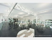 Interno di un ristorante moderno e luminoso con pavimento scuro lucido, tavoli rotondi con sedie bianche e soppalco con ringhiera in vetro. Soffitto alto con travi metalliche bianche a vista e grandi finestre ad arco lungo le pareti.