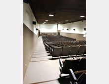 Sala auditorium con file di poltrone disposte in gradinata e corridoi laterali, rivolta verso uno schermo bianco frontale. Pareti bicolori con altoparlanti montati e soffitto scuro con luci a pannello incassate.